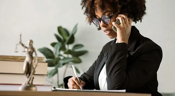 Eine Frau am Telefon notiert etwas auf einem Zettel, daneben steht Justitia in Form einer Miniaturstatue auf dem Tisch.